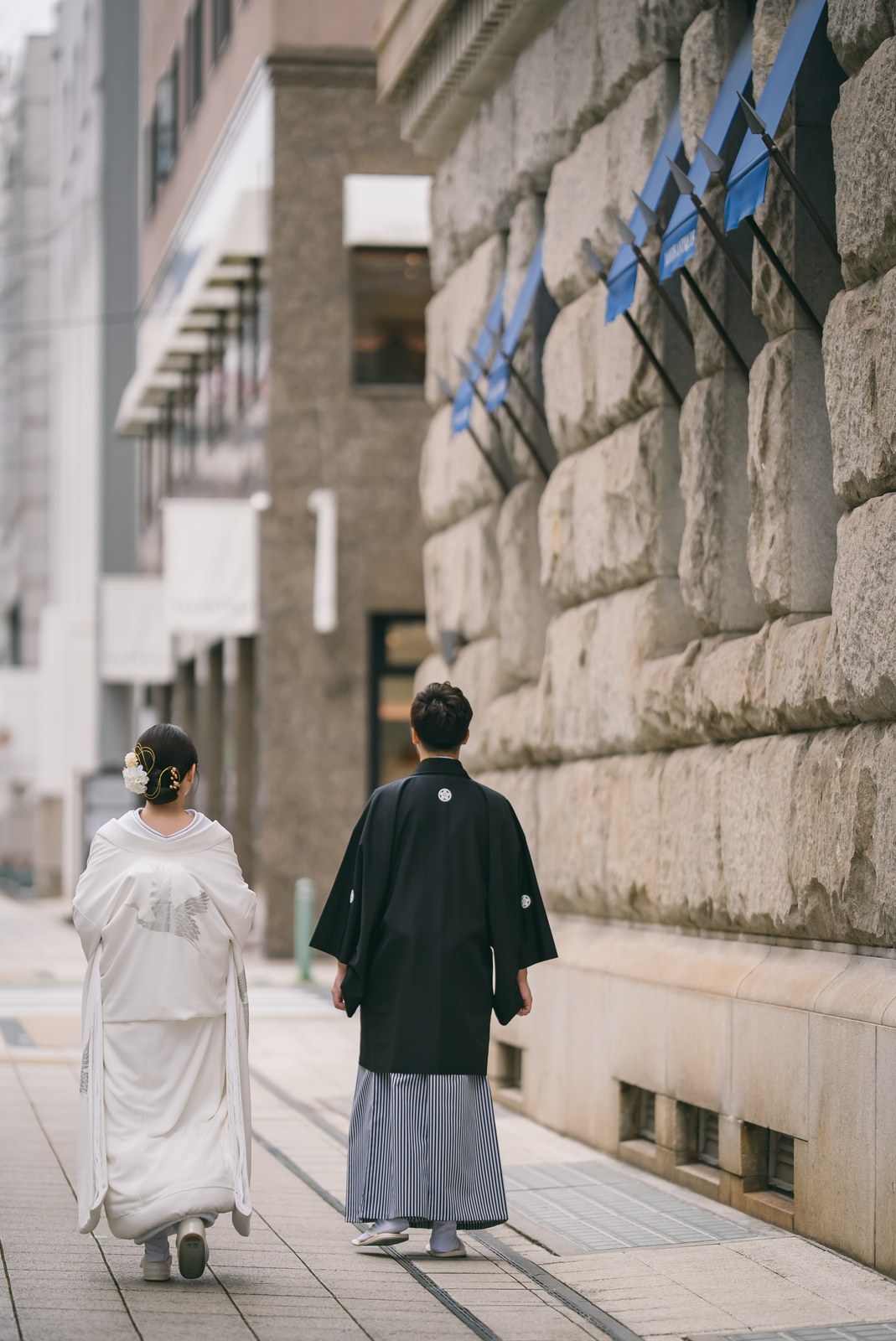 Bride in white shiromuku and groom in black montsuki walking away along a stone-clad building in Kobe