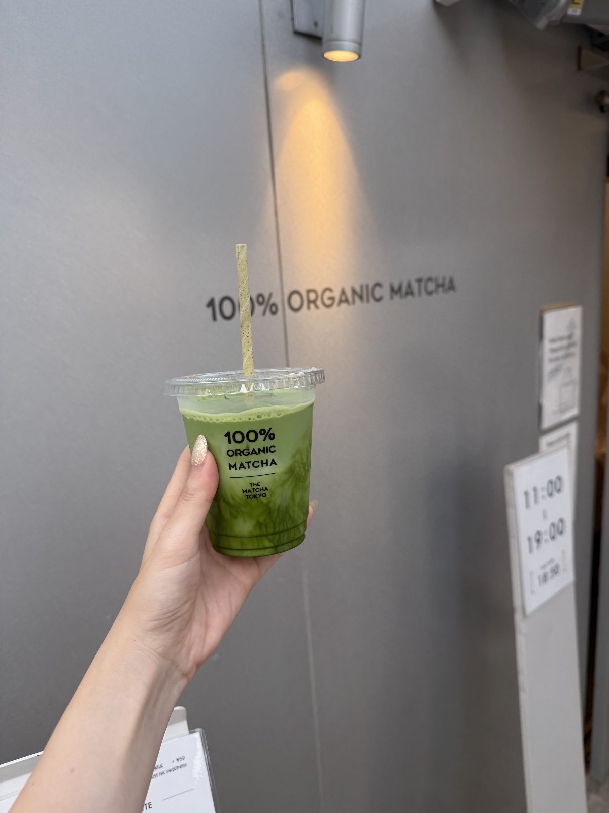 Hand holding an iced Gold Blend matcha cup in front of the 100% Organic Matcha wall at THE MATCHA TOKYO Harajuku