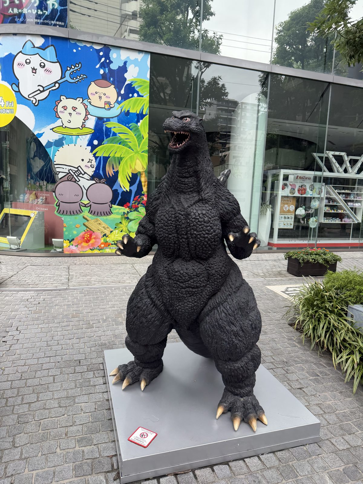 Life-size Godzilla statue on the pavement outside the Bandai headquarters in Komagata Tokyo with a Chiikawa movie mural behind