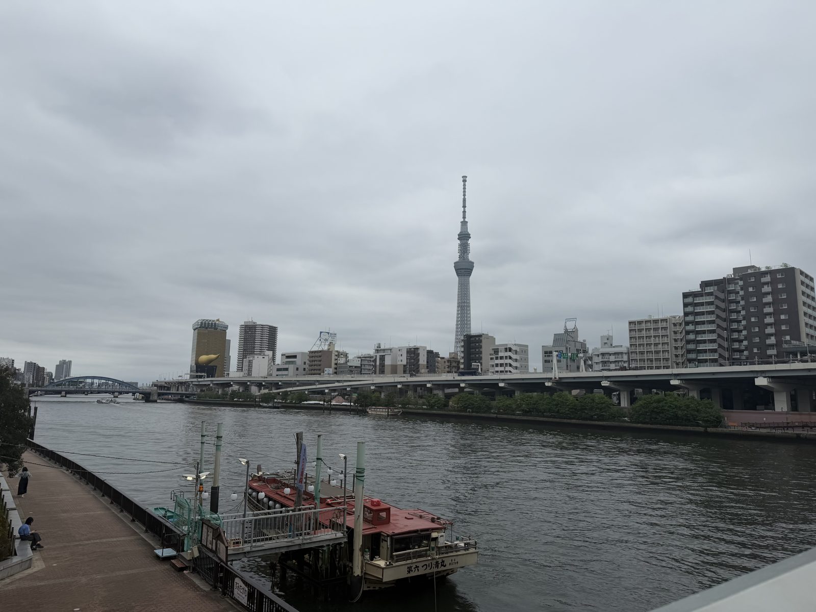 View of the Sumida River from the Kuramae riverside terrace with Tokyo Skytree and the golden Asahi flame under grey skies