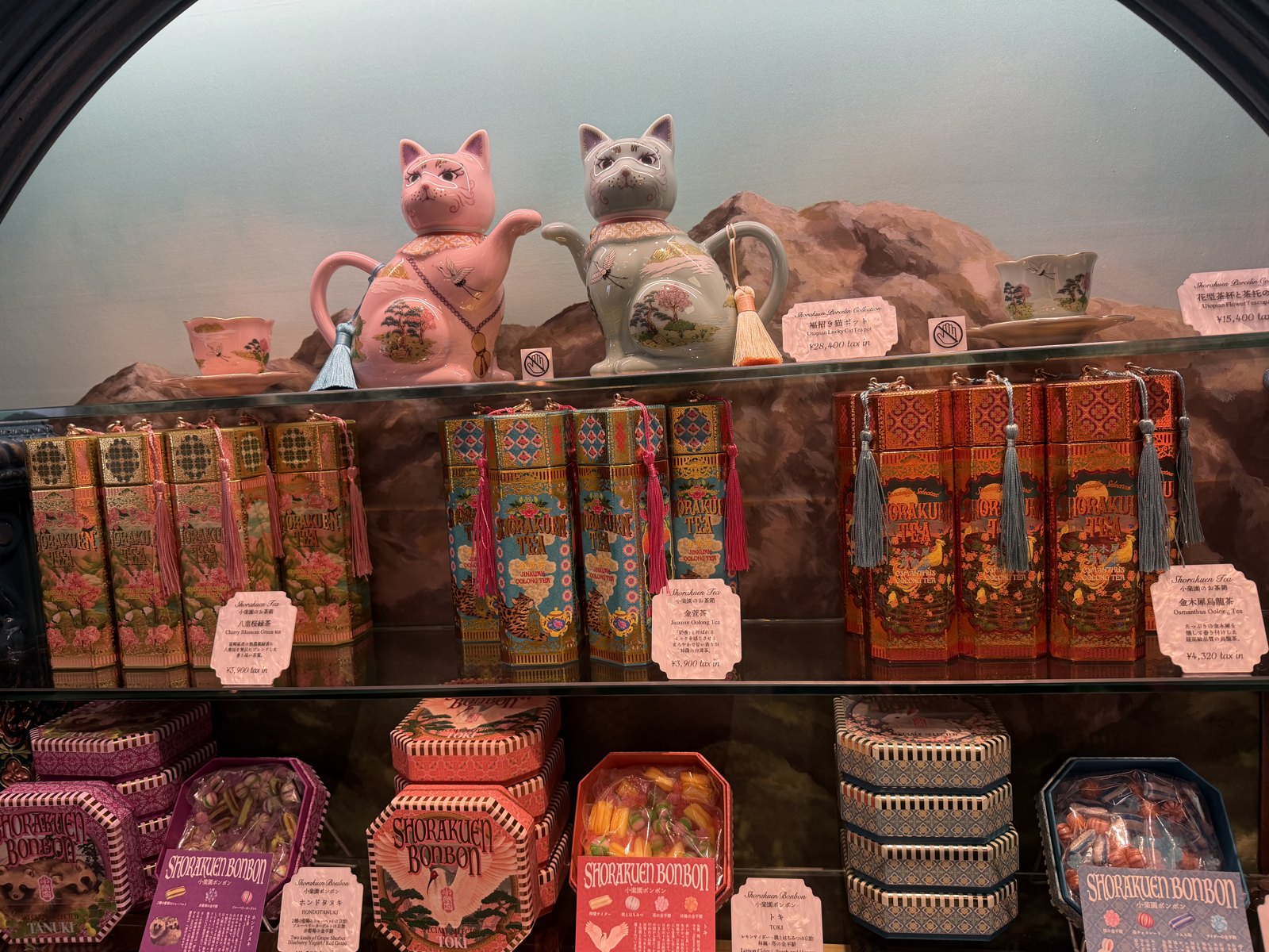 Lucky-cat teapots on a shelf above tall decorated tea tins and octagonal sweet tins